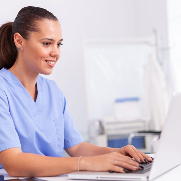 Medical nurse in uniform using laptop sitting at desk in hospital office. Health care physician using computer in modern clinic looking at monitor, medicine, profession, scrubs.